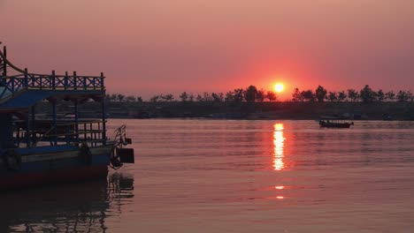 time lapse of beautiful orange sun set in the background over the horizon of the far shore of the lake with a boat coming to park and a parked blue tourist boat bobbing up and down in the water