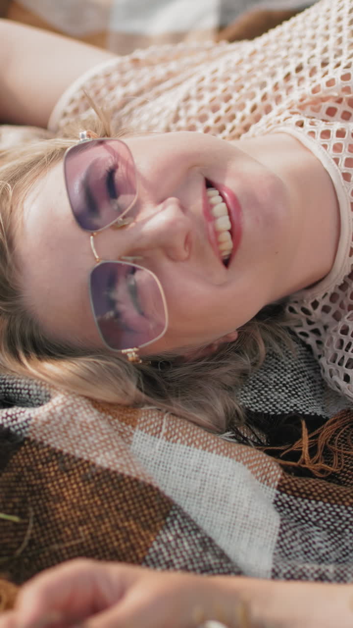 mujer con camiseta y gafas de sol acostada en una manta a cuadros en un campo cubierto de hierba, sonriendo alegremente mientras toca la hierba, un momento relajado al aire libre con la suave luz del sol