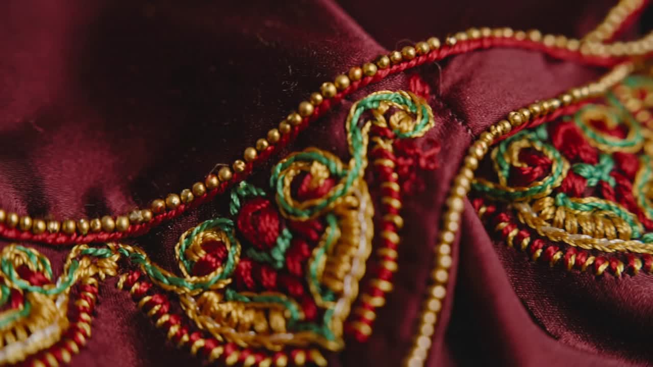 Opening closeup camera panning across maroon textile in studio showing gold beads and green threads