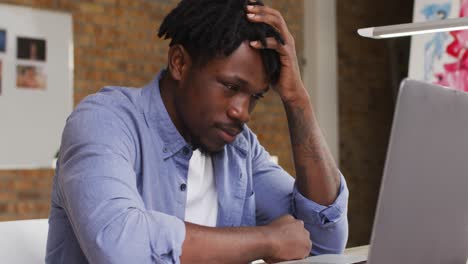 Stressed african american male artist looking at his laptop while sitting on his desk at art studio