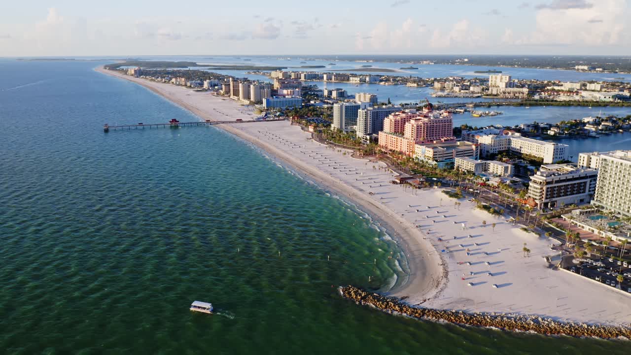 Clearwater Beach curves along the Gulf of Mexico with a pier stretching into the water, as beachfront resorts and umbrellas line the bright sandy shoreline