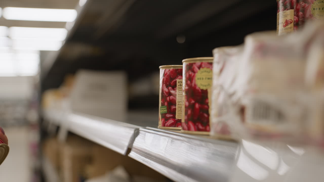 Close up of female shopper hand reaching for can of beans on supermarket shelf wearing smartwatch and white sweater inspect label before grabbing product under fluorescent lights in retail aisle