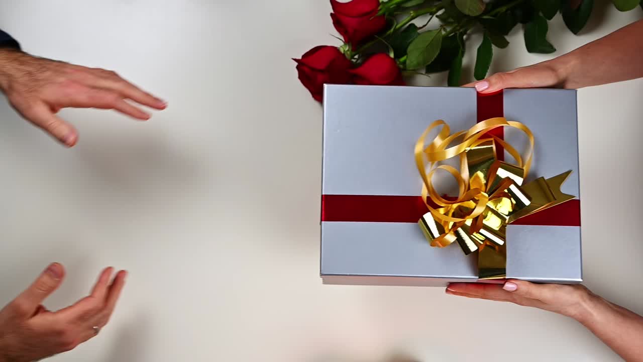 Man's hands give a gray gift box with red and golden ribbons to a woman. Red roses and candles on a white table. Valentines day. Relationships and feelings concept. Overhead shot. Slow motion
