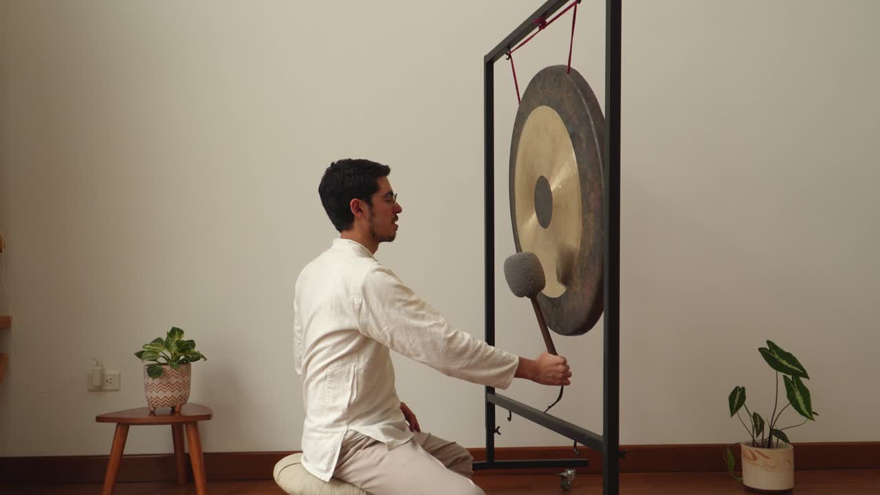 Side-view of a man kneeling on a cushion, holding a soft mallet before a large symphonic gong in a calm minimalist indoor studio with plants—ideal for meditation, wellness and sound healing themes