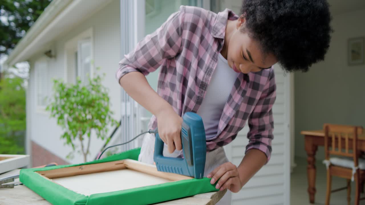 Donna in camicia a quadri rosa che fissa tessuto verde avvolto con una pinzatrice a una cornice sul portico, spazio per il testo.