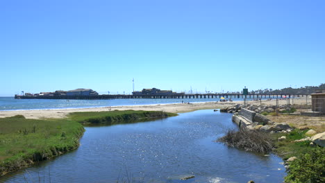 standing on the beach looking over a small lagoon on the beach flowing out into the pacific ocean with stearns wharf pier in the distance on a sunny day in santa barbara, california