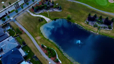 Mansión-De-Ojo-De-Pájaro-De-Sobrevuelo-Aéreo-De-Verano-Junto-Al-Club-De-Golf-Con-Reflejo-Del-Cielo-En-El-Estanque-Artificial-Centrado-Con-Fuentes-Y-Racimos-Simétricos-De-Jardines-Mínimos-Con-Pinos-Rodeados-De-Piedras