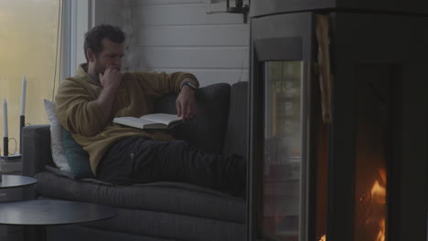 caucasian man sits in sofa reading book with warm fireplace in the foreground, rainy cold autumn outside