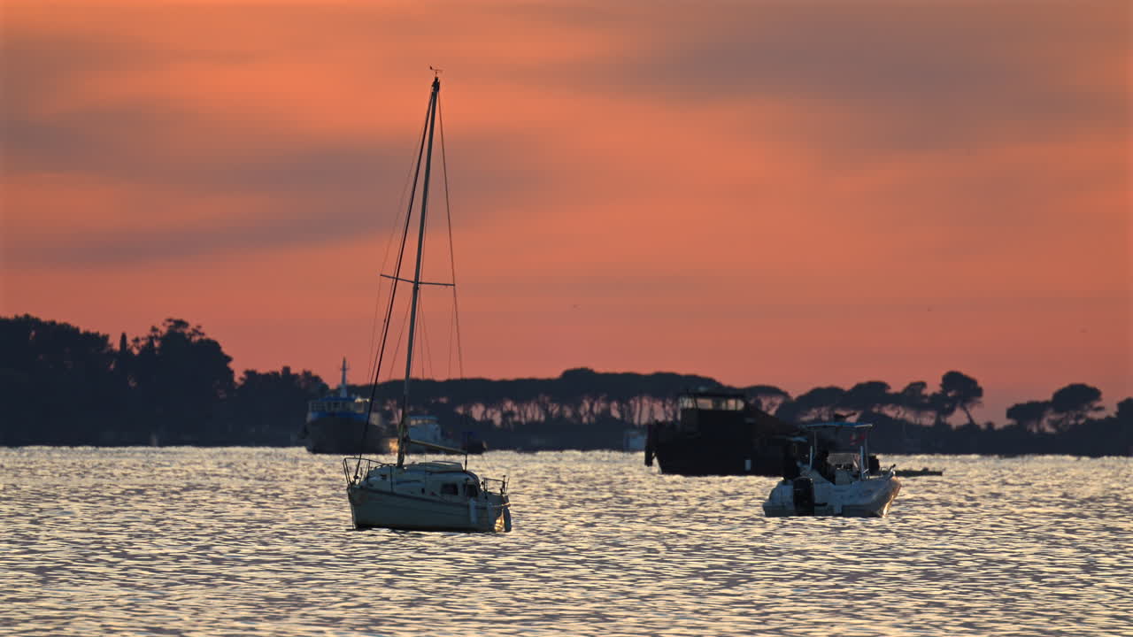 Piccole imbarcazioni ancorate al largo durante il tramonto, con calde tonalità arancioni e rosa che si riflettono sul mare e silhouette di alberi in lontananza