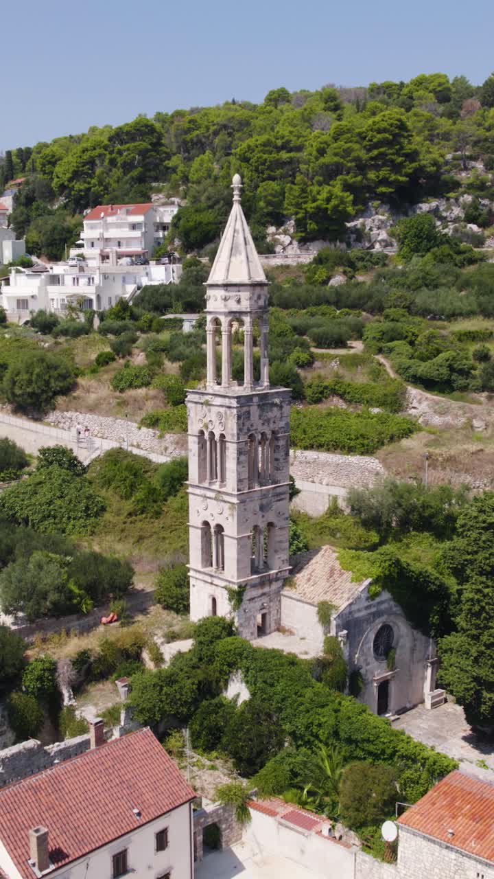 Aerial view of the historic St. Mark's Church bell tower in Hvar, Croatia, surrounded by lush vegetation. Vertical Video, Orbit Motion