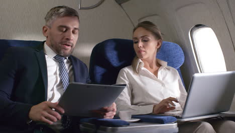 alegre mujer de negocios con laptop y hombre de negocios barbudo sosteniendo portapapeles con documentos discutiendo el trabajo y dándose la mano mientras viaja en avión