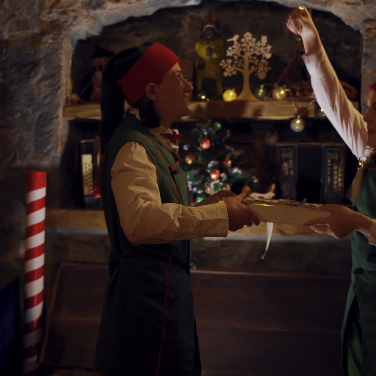 Curious elves in small decorated room opening the magic box. Two elves in fairy suits taking present from the chest in a dark Christmas room.