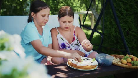 two young girls decorating a cake outdoors on a sunny day with fresh fruits and flowers nearby