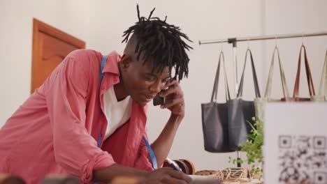 happy african american craftsman with dreadlocks on phone call, taking an order at leather workshop