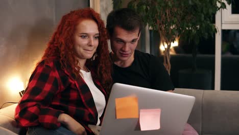 beautiful young couple sitting together with grey laptop on couch. couple - shorthaired man and red headed girl watching together something funny on laptop. talking and smiling. sitting on sofa in stylish loft living room in the evening. close up