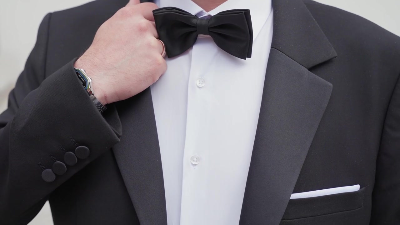 Primer plano detallado de un hombre elegante ajustándose la pajarita negra y la chaqueta, destacando el esmoquin formal, la camisa blanca y los accesorios bajo una luz natural nítida.