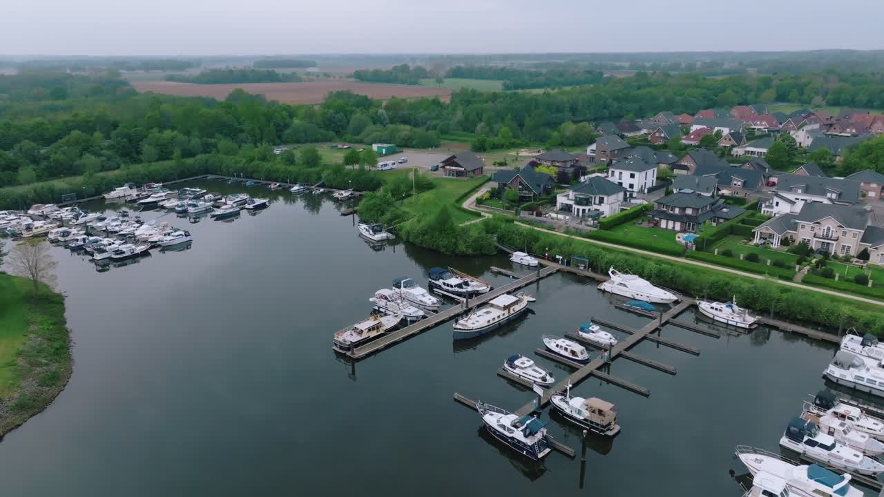 Zoom out revealing boats at the marina and the adjacent residential neighborhood in Doerpen. Soft lighting and cloudy sky define the tranquil scene.