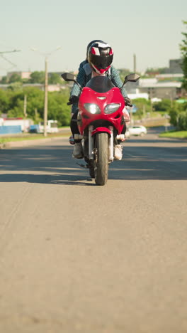 dos amigos, ambos con cascos, viajan en una bicicleta eléctrica roja a lo largo de una carretera de la ciudad, en la distancia, se ve un coche blanco haciendo un giro, con otros vehículos moviéndose en el carril opuesto