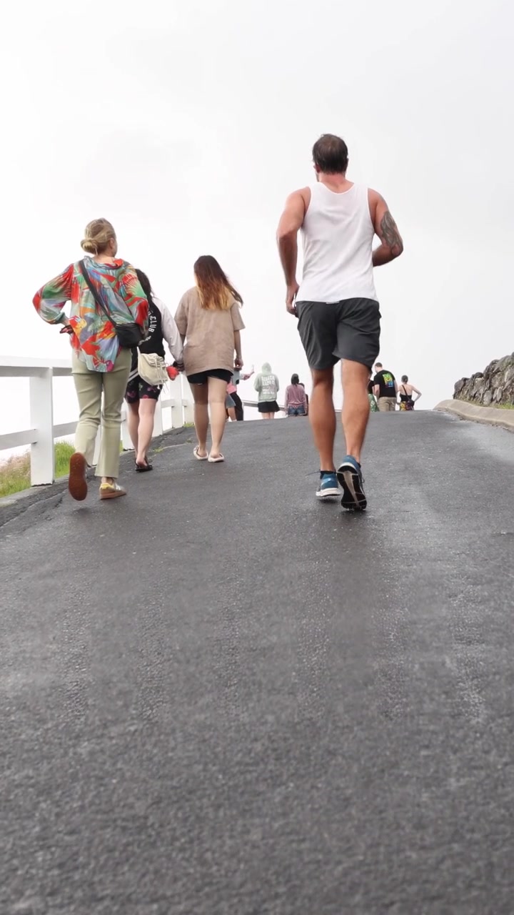 Un grupo de personas camina cuesta arriba por un sendero pavimentado en Byron Bay, Australia. La iluminación nublada y una cámara estable capturan la escena.