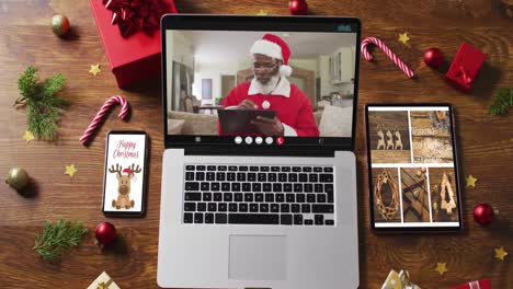 Älterer-Afroamerikanischer-Weihnachtsmann-Bei-Videoanruf-Auf-Laptop,-Mit-Smartphone,-Tablet-Und-Dekorationen