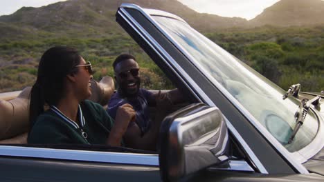 diverse couple driving on sunny day in convertible car raising their arms in the air and smiling