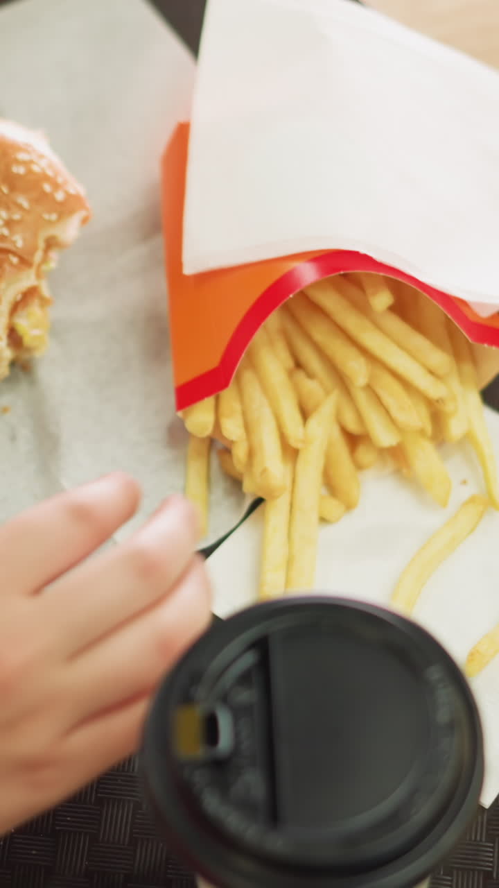 한 여성이 부분적으로 먹은 햄버거를 떨어뜨리는 손으로 트레이에 있는 킨에 오른손을 니다. 트레이에는 감자김과 나무 테이블에 있는 커피 컵이 있습니다. 그녀는 김을 가져와 한 어리를 먹습니다.