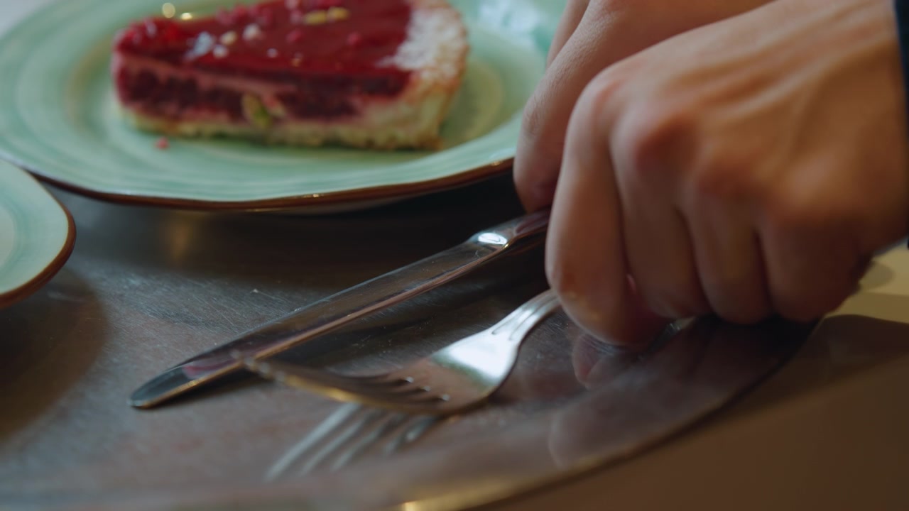 Nærbillede af en hånd, der rækker ud efter bestik på et bord med tallerkener og mad. En kniv hviler ved siden af en gaffel, mens hænder griber redskaber over grønne keramiske skåle med desserter. Blød belysning.