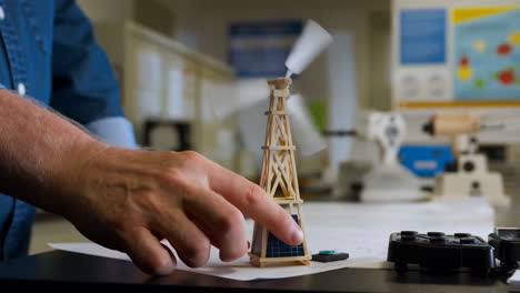a person's hand next to a miniature windmill model with solar panels in a lab