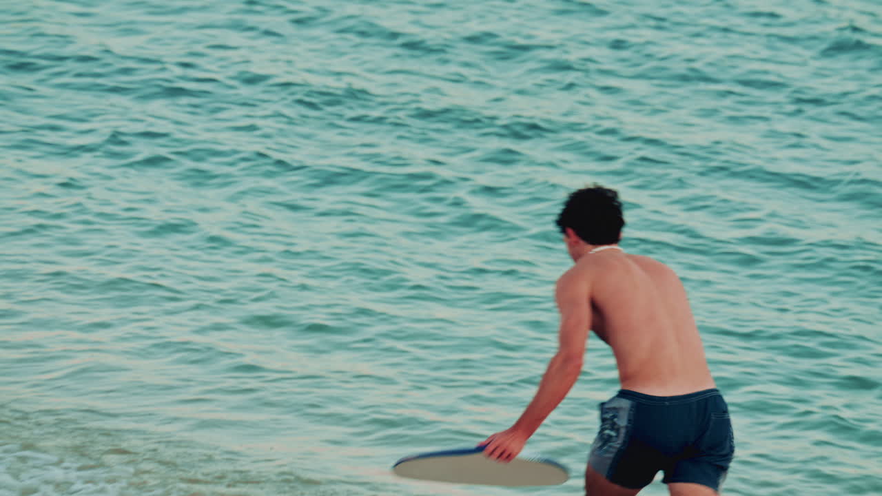 A young man skimboards along the shallow shoreline, balancing as water splashes around him