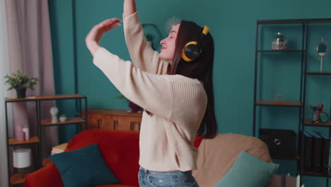 overjoyed young woman in wireless headphones dancing, singing on cozy couch in living room at home