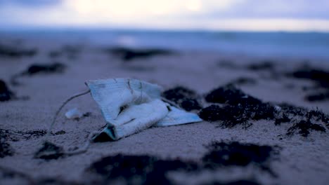 used face mask, trash and waste litter on an empty baltic sea white sand beach, environmental pollution problem, overcast evening after sunset, closeup shot