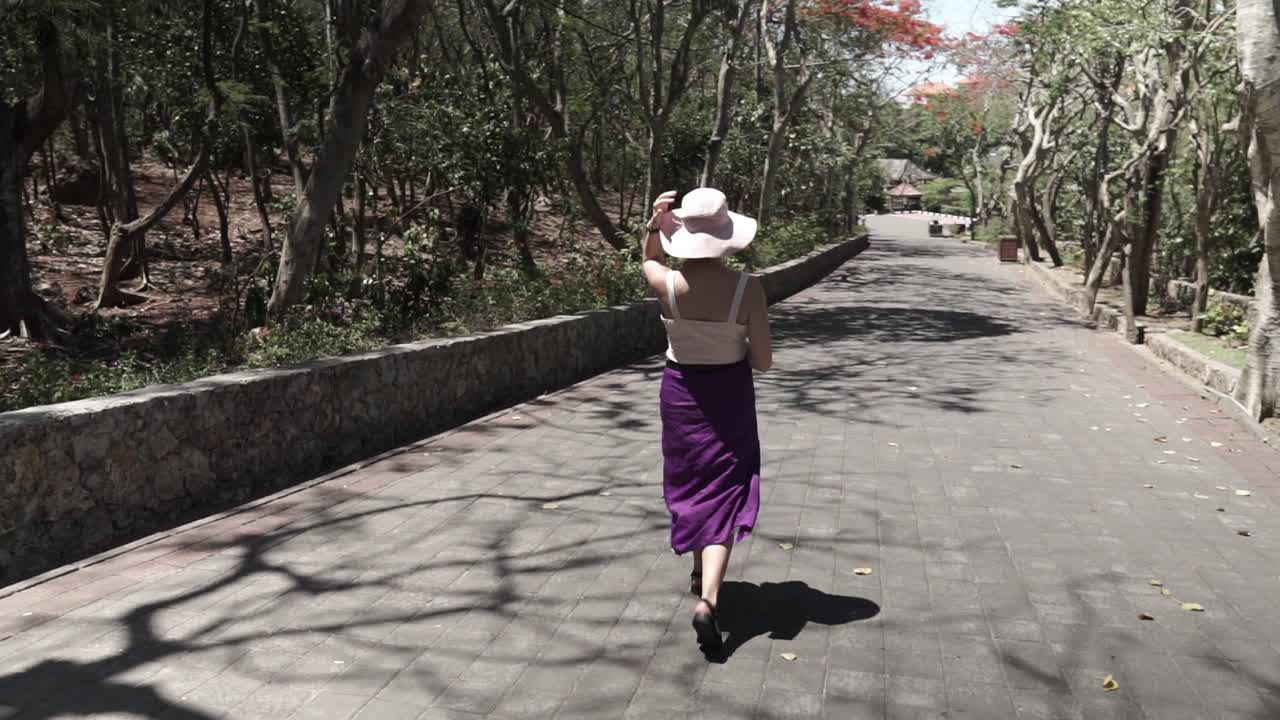 joven turista con un sombrero blanco y una larga falda violeta caminando hacia el parque, indonesia, la cámara sigue y el paralaje