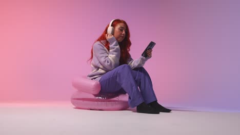 studio shot of young gen z woman wearing headphones streaming music to mobile phone