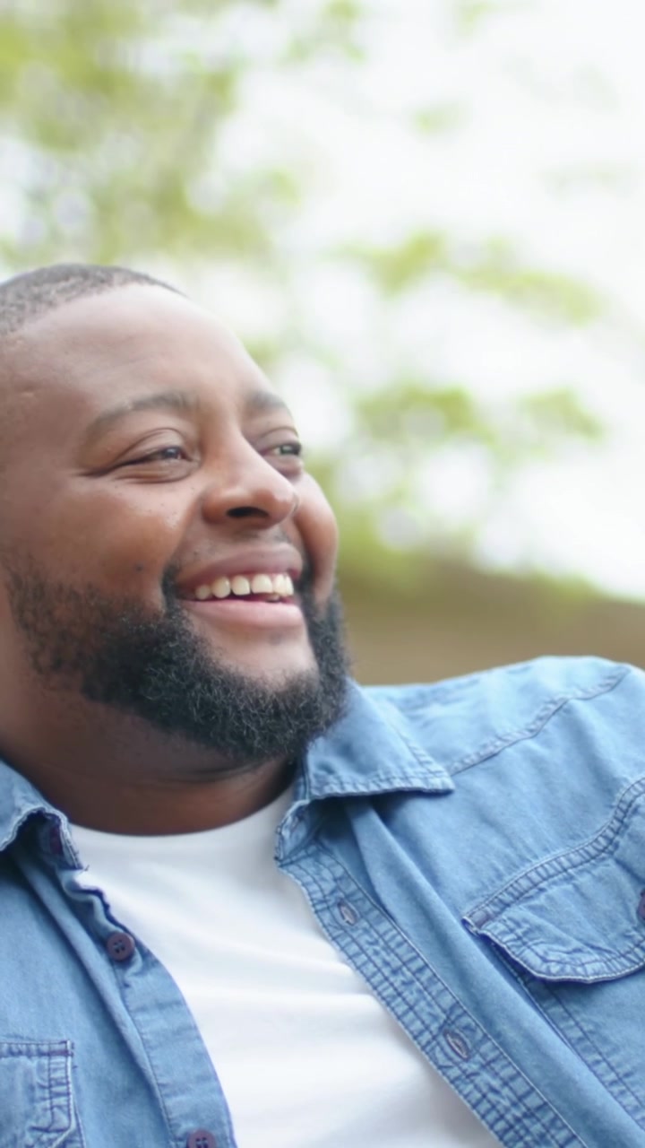 Vertical video: Smiling man laughing as camera rises in park, with denim shirt, white tee and watch