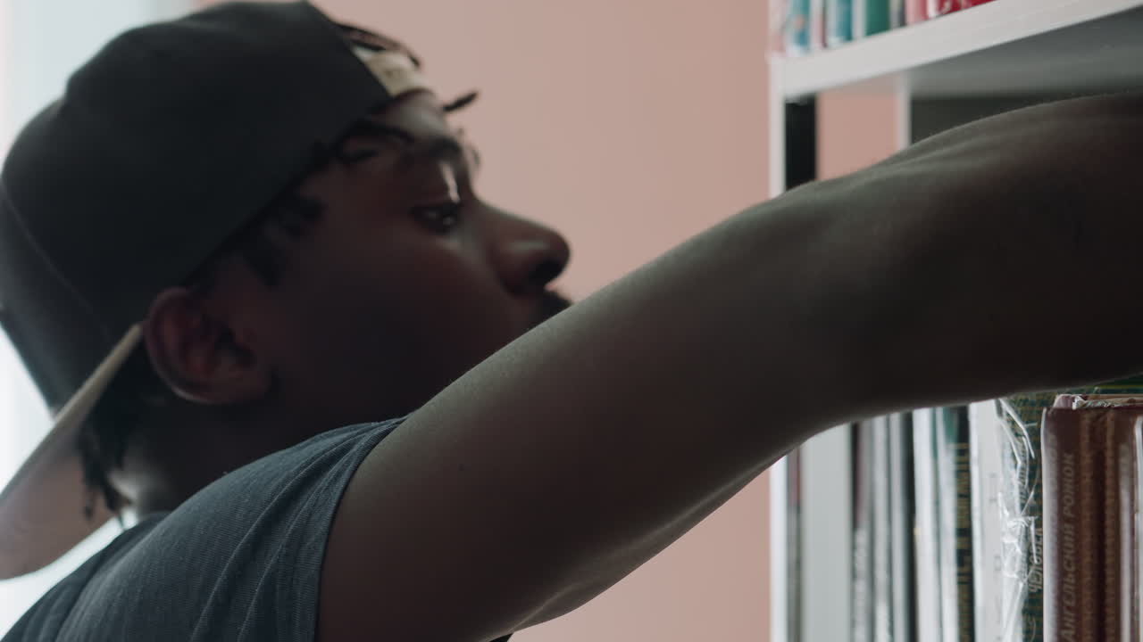 Close up of young man wearing backward cap reaching toward bookshelf selecting book with focused expression in softly lit library environment