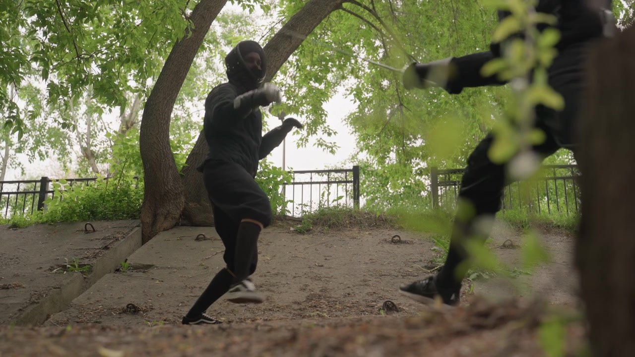 Two people in black fencing gear engage with swords outdoors under trees demonstrating techniques. One person holds sword while other moves into position both wear protective masks