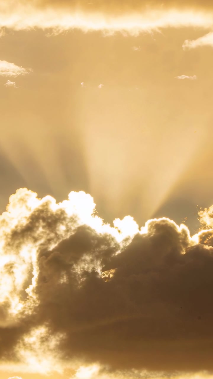 Vertical video: Sun peeking behind dense cloud bank, sending golden sunbeams upward near horizon