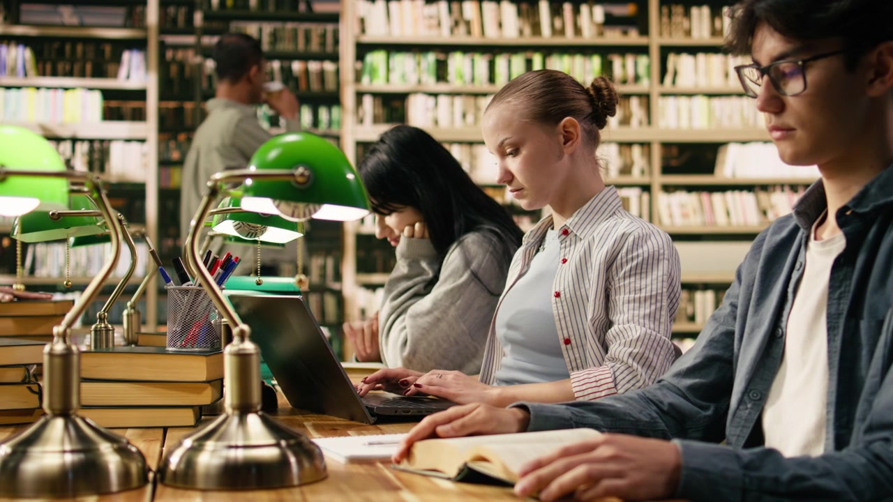 Studente che digita sul laptop e prende appunti da un libro aperto alla scrivania della biblioteca.
