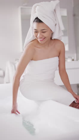 Vertical video of smiling biracial woman with towel on hair sitting on bathtub in bathroom
