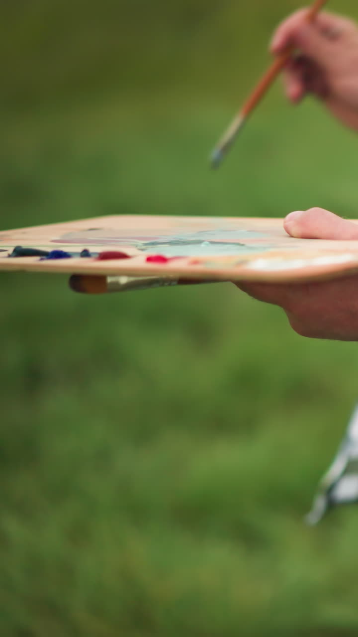 A close-up shot of an artist wearing a checkered shirt, holding a palette full of vibrant paint colors in a serene grassland. The artist's face is not visible