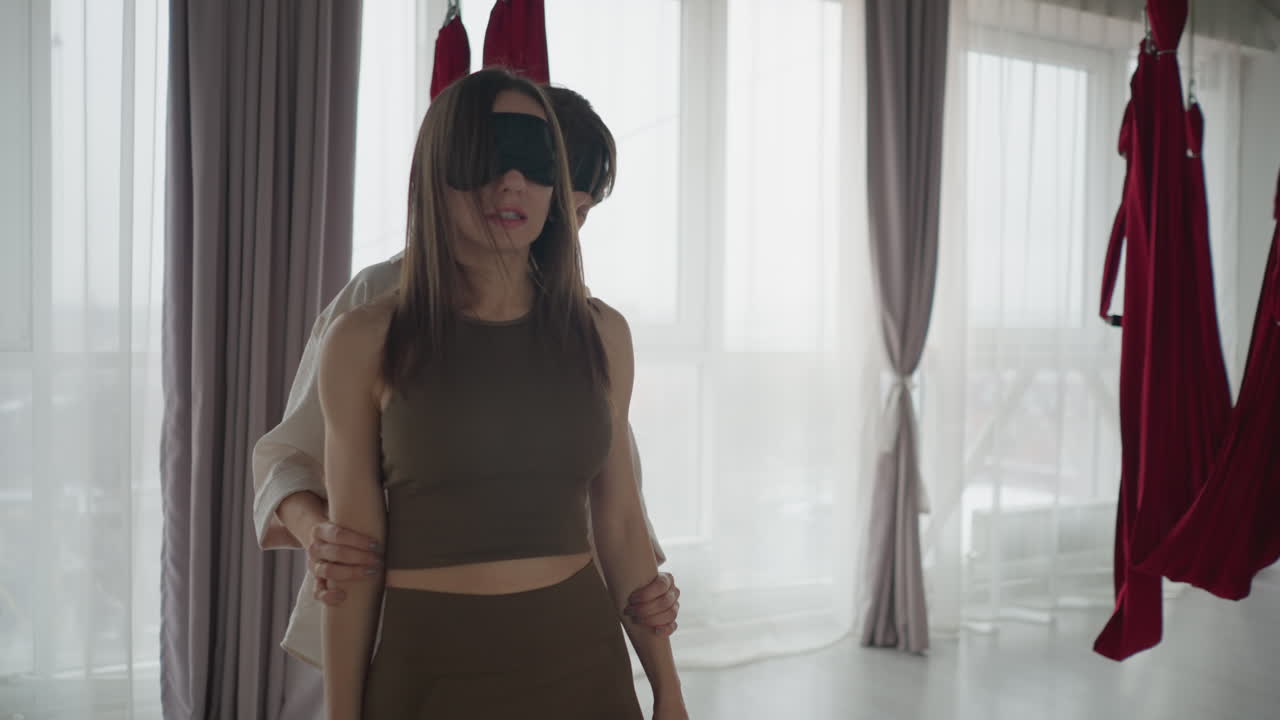 Fitness coach gently holds partner with eyes covered while assisting body stretch in bright studio with red hammocks, showing calm connection and support in mindful movement practice