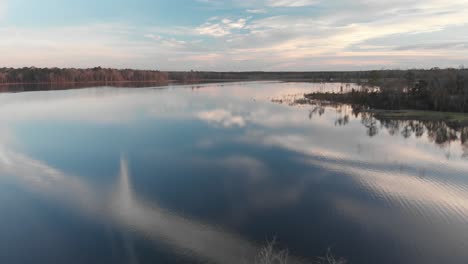 slowly flying backwards over a beautiful lake with the blue evening sky reflected in the gentle ripples of the water, as the setting sun casts a golden glow on the distant trees