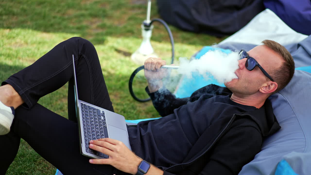 Chilling young man lying in the comfortable bean bag chair. Male in dark glasses smoking shisha and watching video on laptop.
