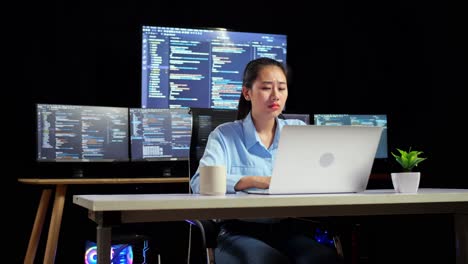 asian female programmer yawning while writing code by a laptop using multiple monitors showing database on terminal window desktops in the office