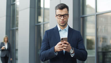 businessman in glases texting and chatting with smartphone in the street