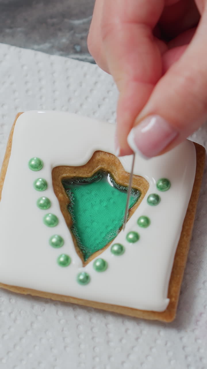 primer plano de manos ajustando cuentas decorativas verdes en una galleta helada con glaseado blanco y relleno verde, la galleta se coloca en un papel de pañuelo con detalles intrincados de la colocación de cuentas para la decoración