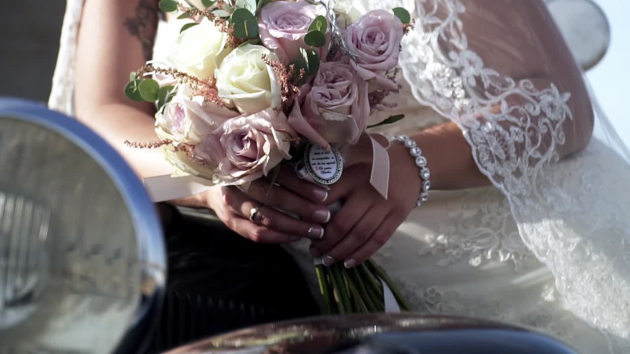 toma manual en cámara lenta de la novia vestida con un vestido de novia blanco y joyas en la mano apoyada en un camión vintage negro con un ramo de rosas en la mano el día de la boda