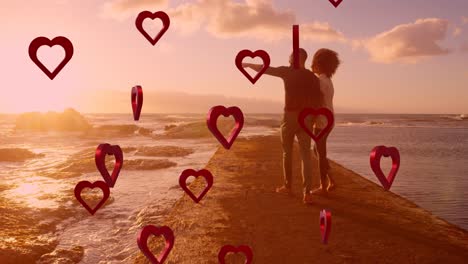 animación de corazones rojos sobre una pareja enamorada tomados de la mano por el mar.