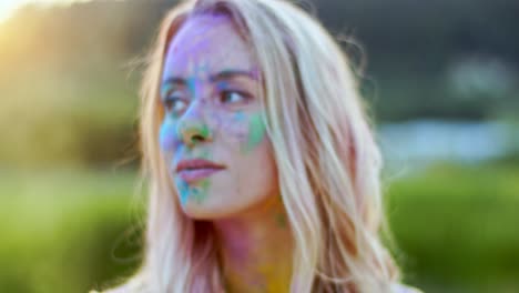 portrait shot of the pretty charming blond woman with colorful paints on her face smiling cheerfully to the camera outdoor at the holi festival