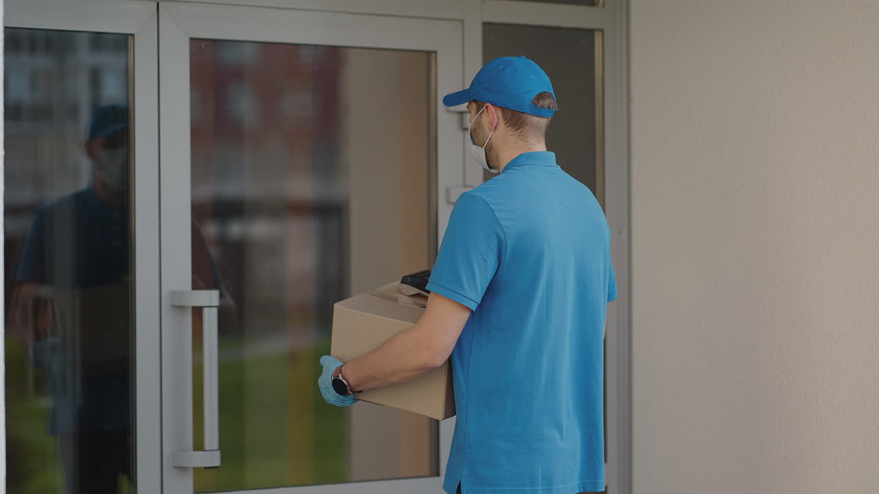Pandemic and online order delivery A delivery man wearing a protective mask and gloves passes the parcel to the customer and receives a contactless payment via tas